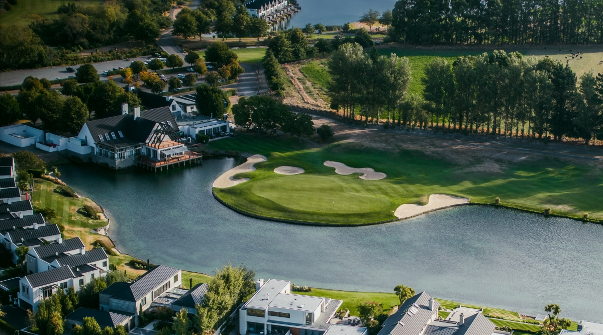The 18th hole at Clearwater Golf Club Christchurch showing the final approach along the sweeping par-4 to the clubhouse with the largest spring-fed lake on the left and the resort facilities visible ahead