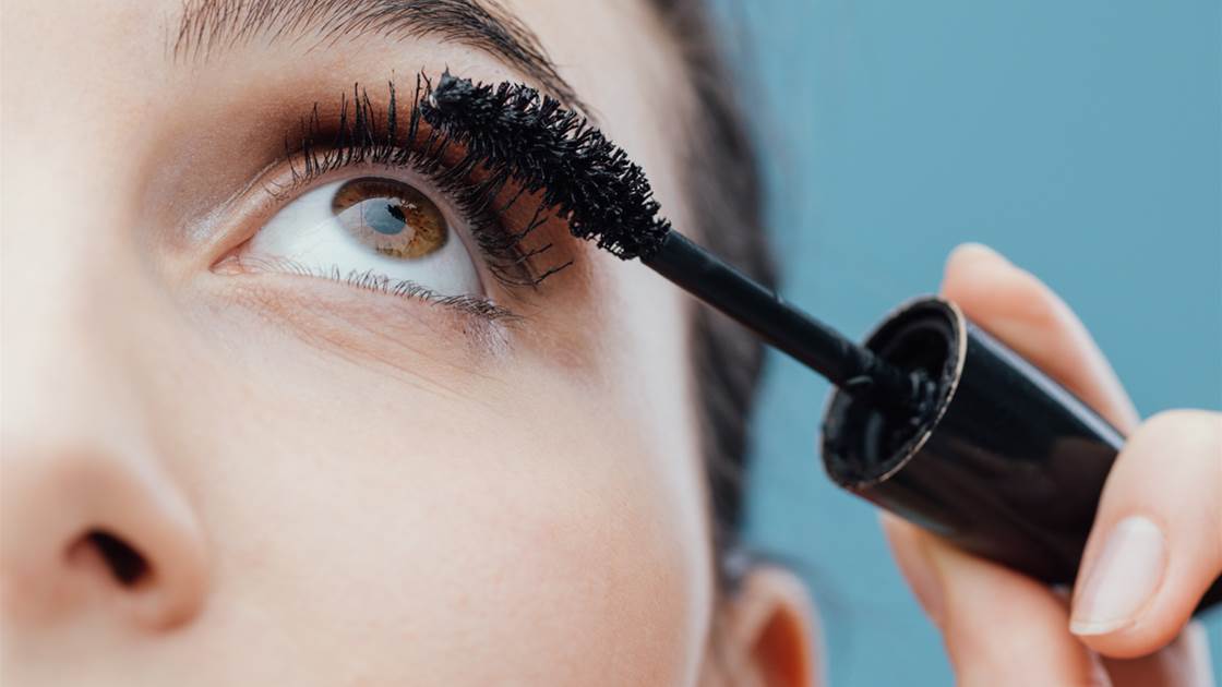 Close-up of woman applying mascara to upper lashes against blue background Close-up of woman applying mascara to upper lashes against blue background