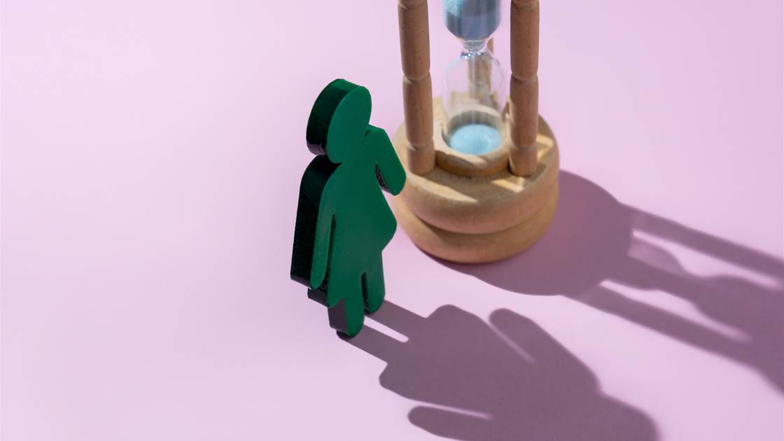 Menopause concept: green female figure beside an hourglass casting a long shadow on a pink background Menopause concept: green female figure beside an hourglass casting a long shadow on a pink background